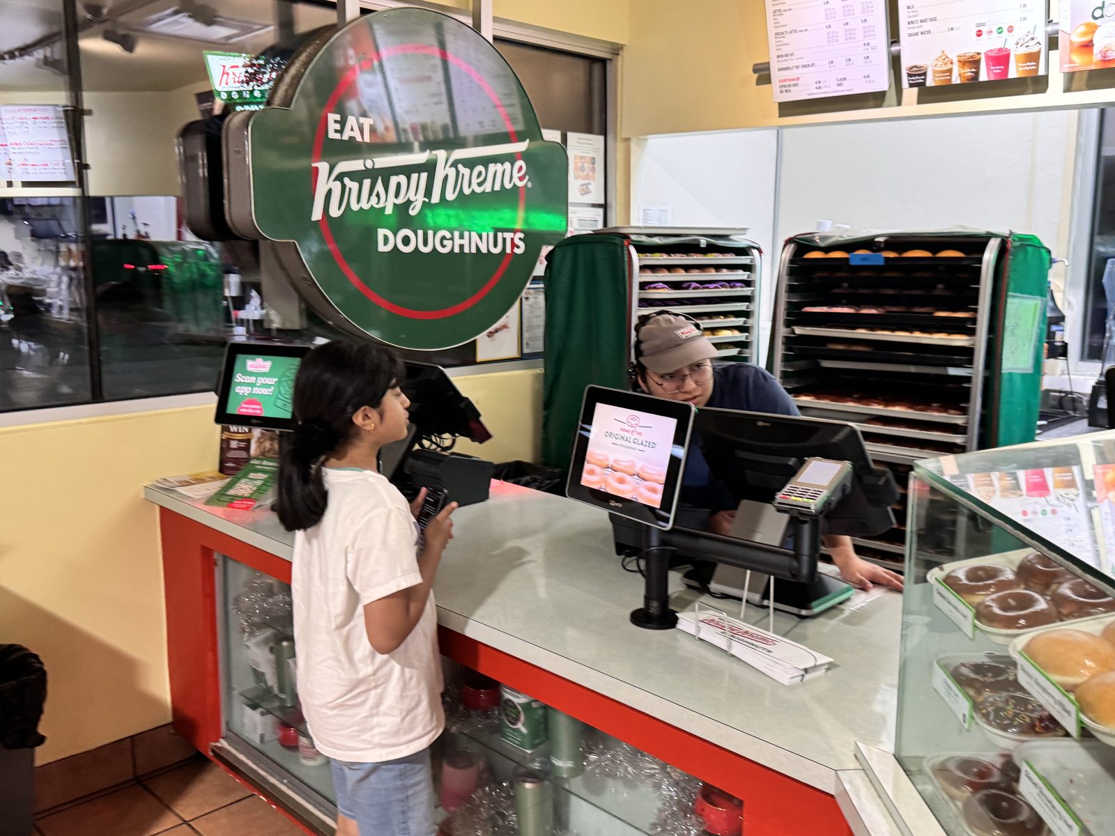 Sri at the Krispy Kreme counter talking to a worker, with empty donut racks visible behind